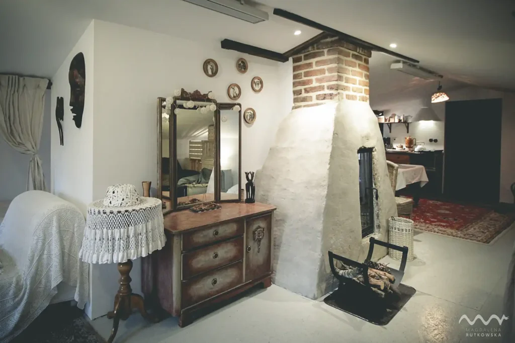 Poddasze. Przytulne wnętrze Poddasza Łuby Siedem. Centralnie umieszczony kominek w glinianej babie, obok zabytkowa toaletka, wiklinowe meble oraz drewniane belki konstrukcyjne.
