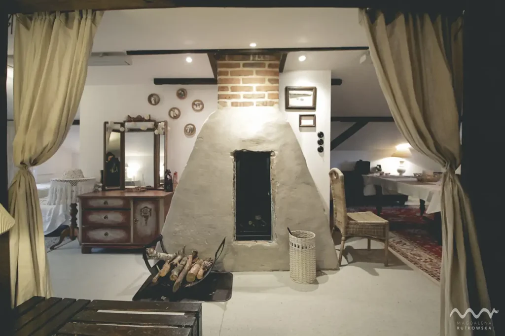 Poddasze, wnętrze. Przytulne wnętrze Poddasza w Łuby Siedem. Widoczny kominek w glinianej babie, zabytkowa toaletka, wiklinowe meble i drewniane belki konstrukcyjne.