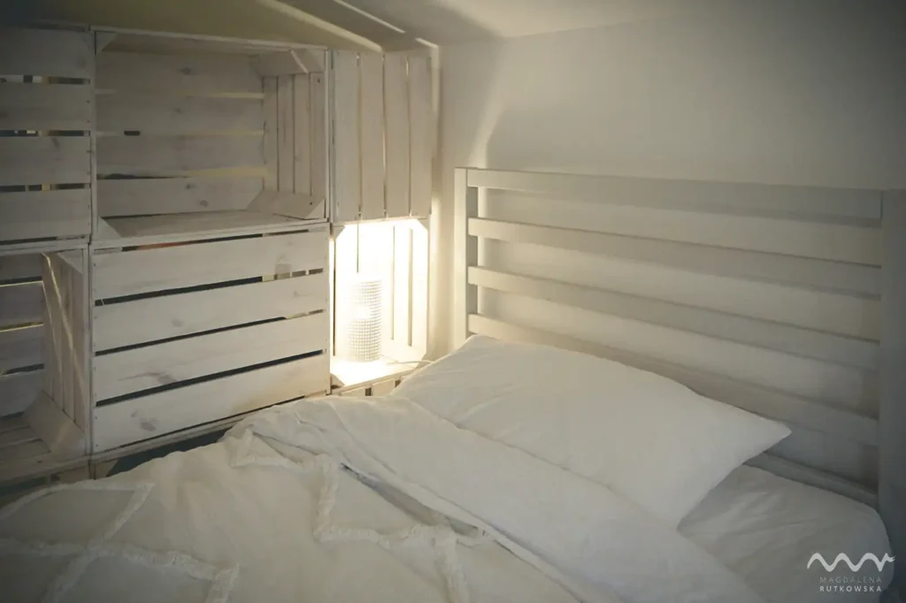 Poddasze Sen. Przytulne łóżko z jasną pościelą na Poddaszu Łuby Siedem, na tle ażurowej ścianki. Włączona lampka nocna zaprasza do wieczornego wypoczynku i slow life.