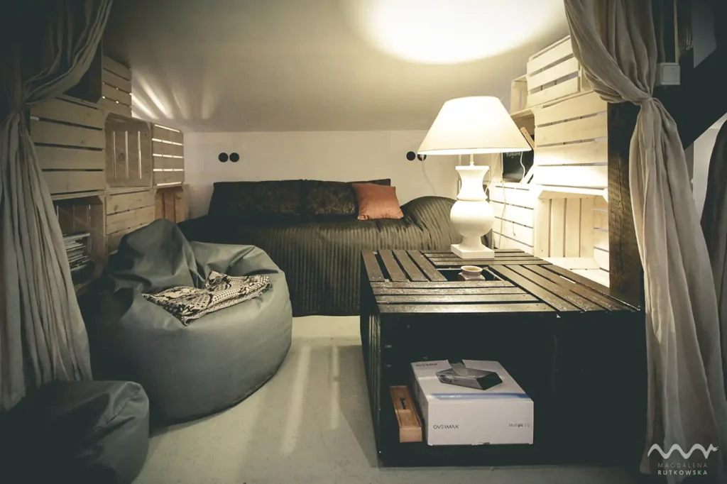 Poddasze. Detal na Poddaszu Łuby Siedem: Jasna lampa na stoliku ze skrzynek, worek sako i łóżko z brązową narzutą. Naturalne lniane zasłony oddzielają przytulną alkowę.