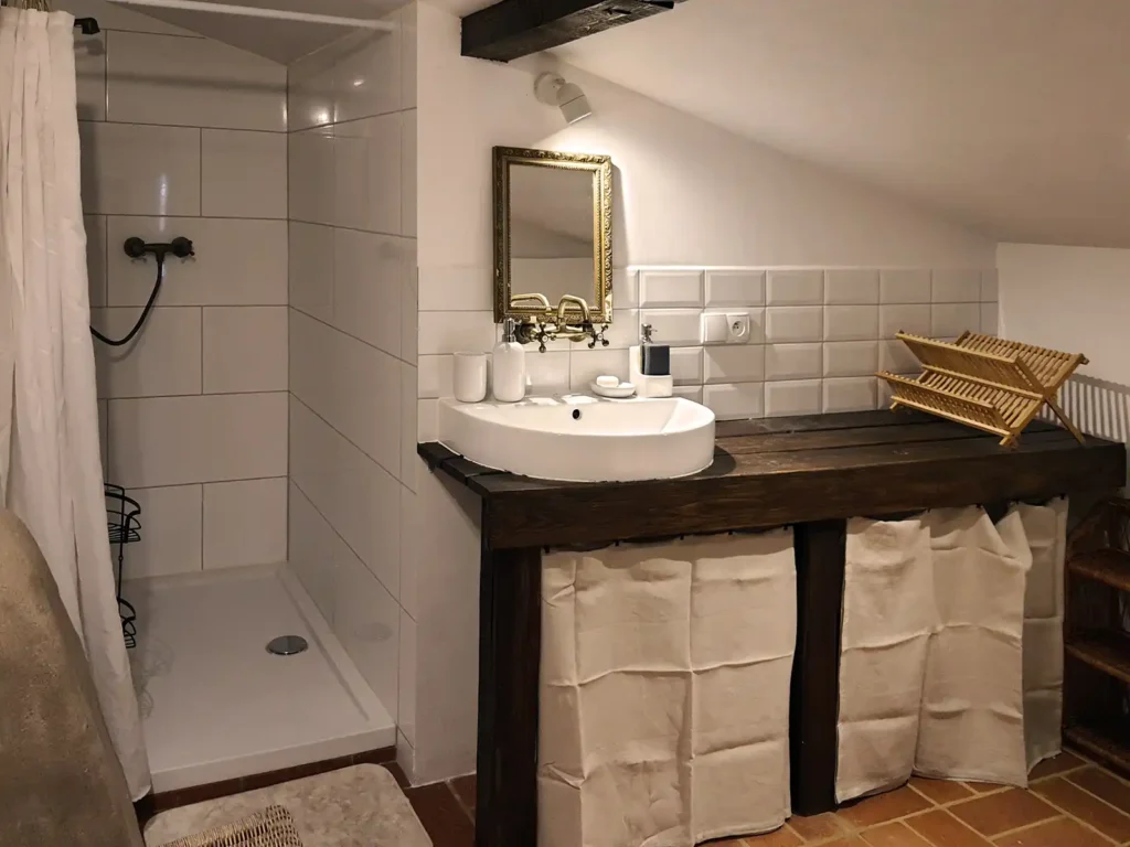Poddasze, łazienka. Łazienka na Poddaszu Łuby Siedem. Widoczny prysznic walk-in, podłoga z czerwonej cegły, drewniany blat, mosiężny kran i gliniana ścianka kominka.