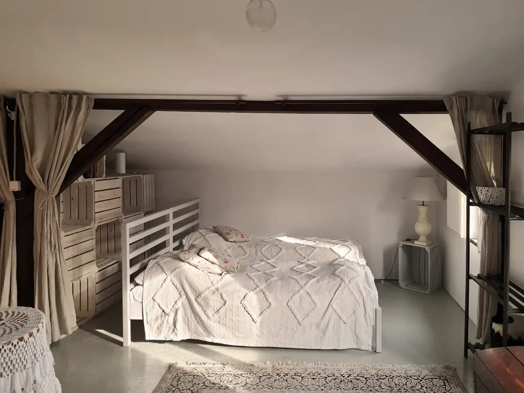 Poddasze. Alkowa na końcu Poddasza Łuby Siedem przy oknie. Dwa łóżka z jasnymi narzutami, oddzielone ażurową ścianką i lnianymi zasłonami, zapewniające intymny nocleg w Borach Tucholskich.