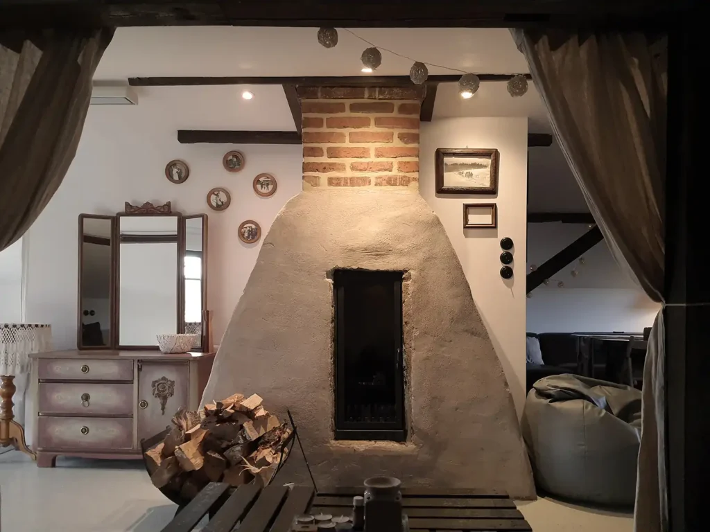 Poddasze. Wnętrze Poddasza Łuby Siedem: kominek w glinianej babie, drewniane belki konstrukcyjne i zabytkowa toaletka, sprzyjające slow life w Borach Tucholskich.