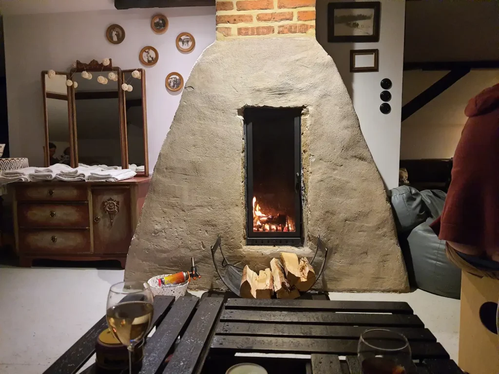 Poddasze, relaks. Wnętrze Poddasza Łuby Siedem: kominek w glinianej babie z płonącym ogniem, kieliszek wina, drewniane belki konstrukcyjne i zabytkowa toaletka, sprzyjające slow life w Borach Tucholskich.