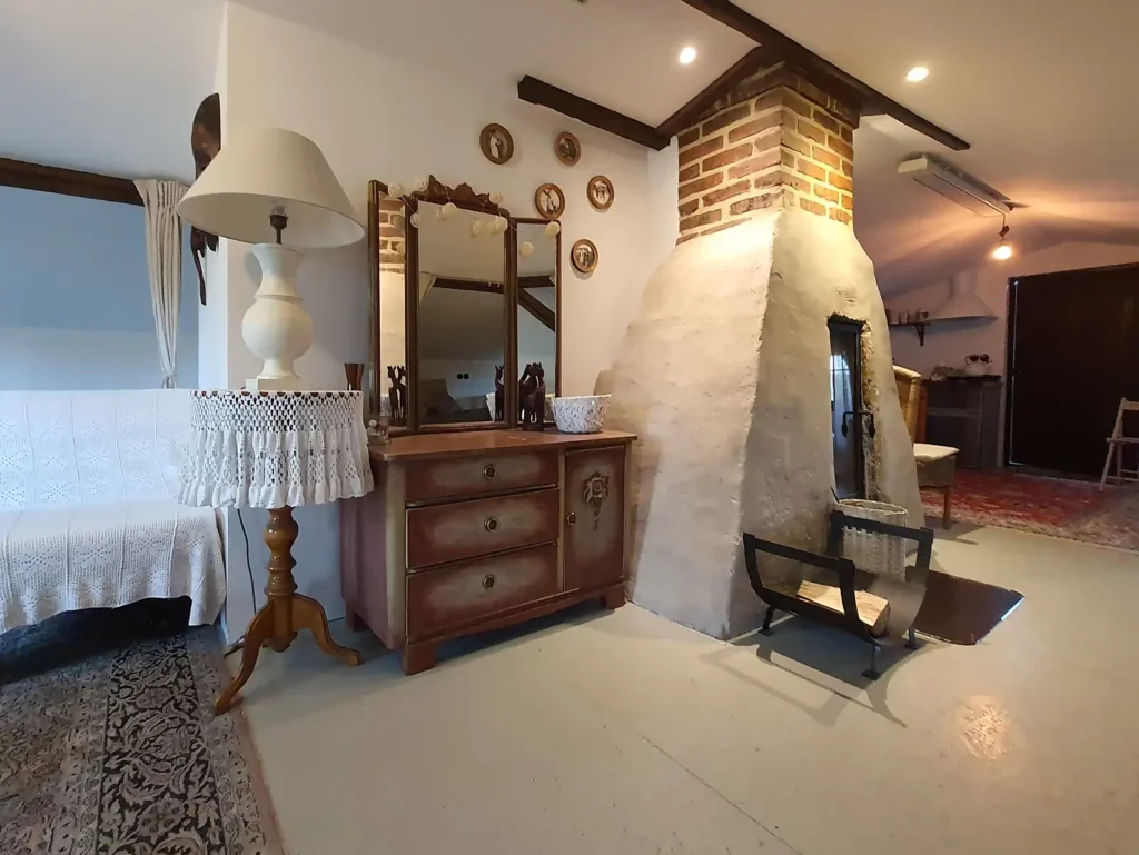 Poddasze. Przytulne wnętrze Poddasza Łuby Siedem. Centralnie umieszczony kominek w glinianej babie, obok zabytkowa toaletka, wiklinowe meble oraz drewniane belki konstrukcyjne.