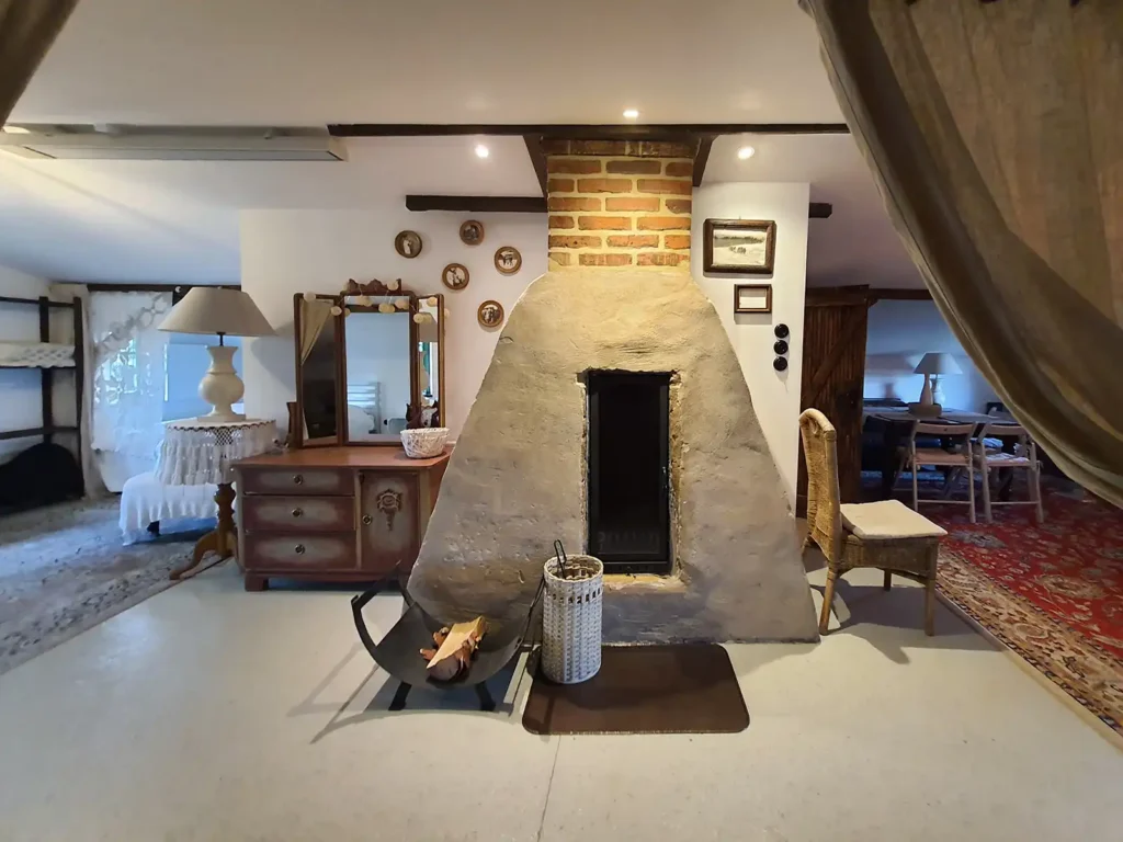 Poddasze, kominek. Przytulne wnętrze Poddasza w Łuby Siedem. Widoczny kominek w glinianej babie, zabytkowa toaletka, wiklinowe meble i drewniane belki konstrukcyjne.