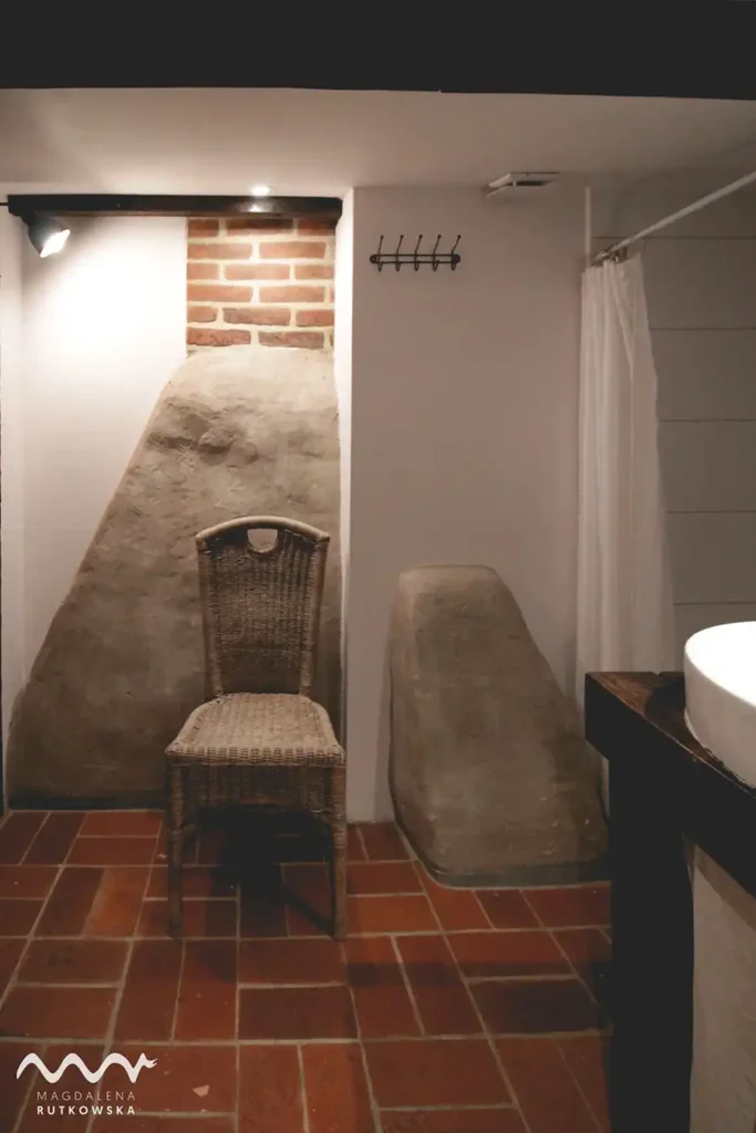 Poddasze, łazienka. Łazienka na Poddaszu Łuby Siedem. Podłoga z czerwonej cegły, drewniane belki, gliniana ścianka kominka baby i wiklinowe krzesło.