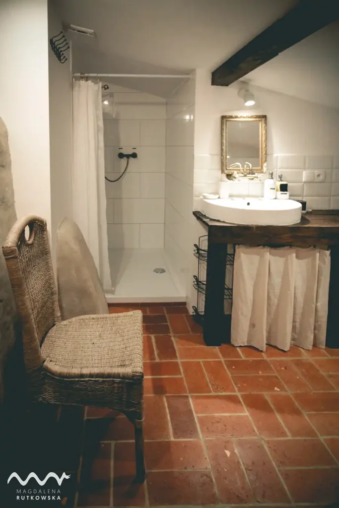 Poddasze, łazienka. Łazienka na Poddaszu w Łuby Siedem. Widoczny prysznic walk-in, podłoga z czerwonej cegły, drewniany blat, mosiężny kran i fragment glinianej ścianki kominka.