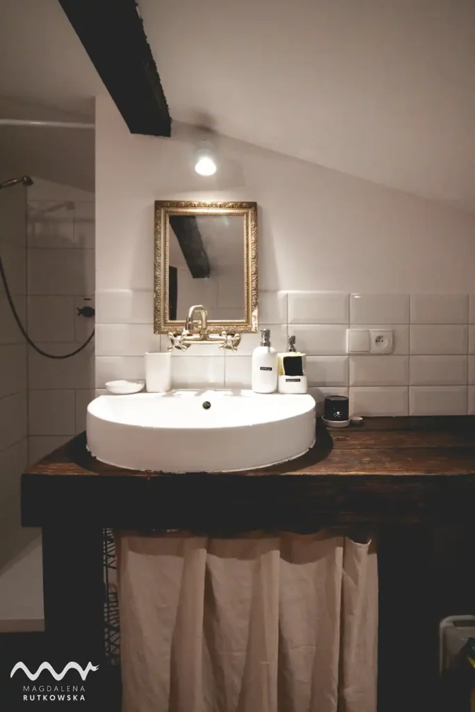 Poddasze, łazienka. Wąski kadr łazienki na Poddaszu Łuby Siedem z umywalką nablatową, drewnianym blatem, mosiężnym kranem i ozdobnym, zabytkowym lustrem.