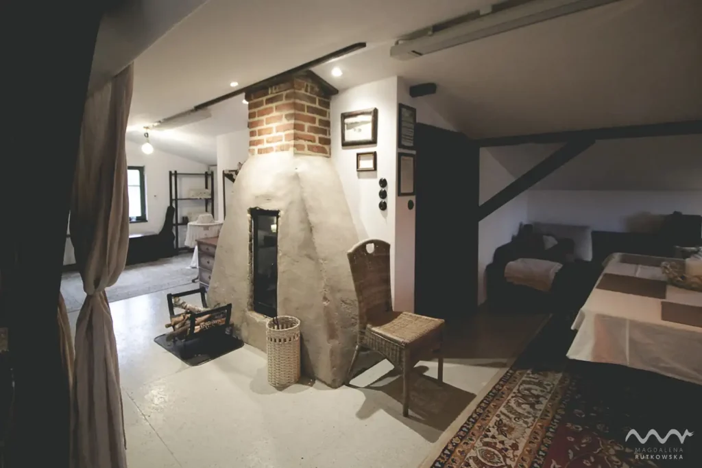 Poddasze. Kominek z glinianą babą i wiklinowe meble na Poddaszu w Łuby Siedem. Widać zasłonę lnianą oddzielającą łóżko oraz małe okna z zielonymi framugami.