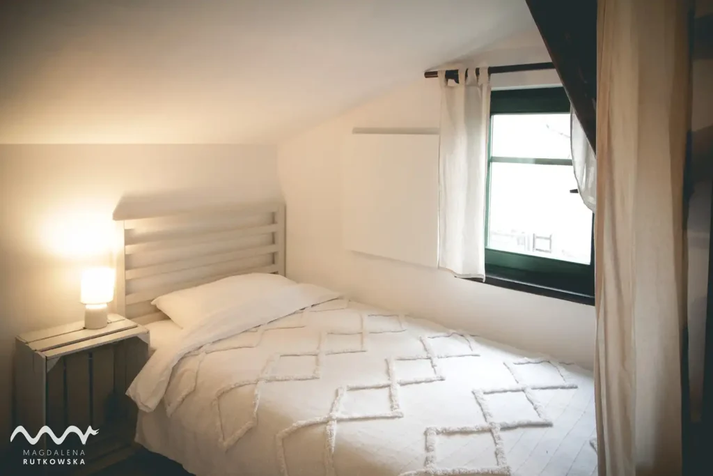 Poddasze. Łóżko z jasną narzutą i lampką nocną na drewnianym stoliku ze skrzynki na Poddaszu Łuby Siedem, Oddzielone lnianą zasłoną. Widać drewniane okno z zieloną framugą.