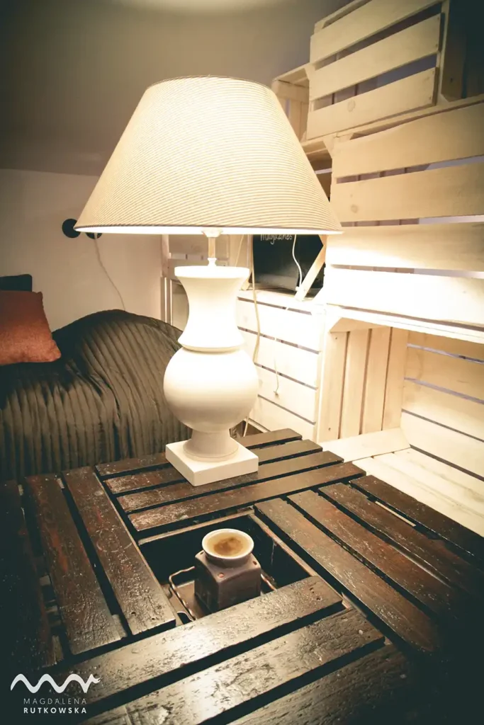 Poddasze. Detal na Poddaszu Łuby Siedem: Jasna lampa stołowa z płóciennym abażurem stoi na stoliku z drewnianych skrzynek, w tle ścianka z drewnianych skrzynek. Kominek na olejki eteryczne.
