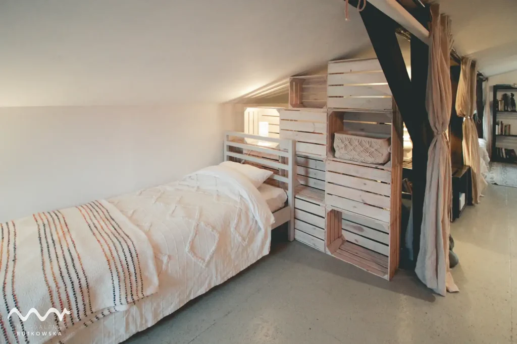 Poddasze, nocleg. Łóżko z jasną narzutą na Poddaszu Łuby Siedem, oddzielone lnianą zasłoną, na tle ażurowej ścianki z drewnianych skrzynek. Widać drewniane belki stropowe.