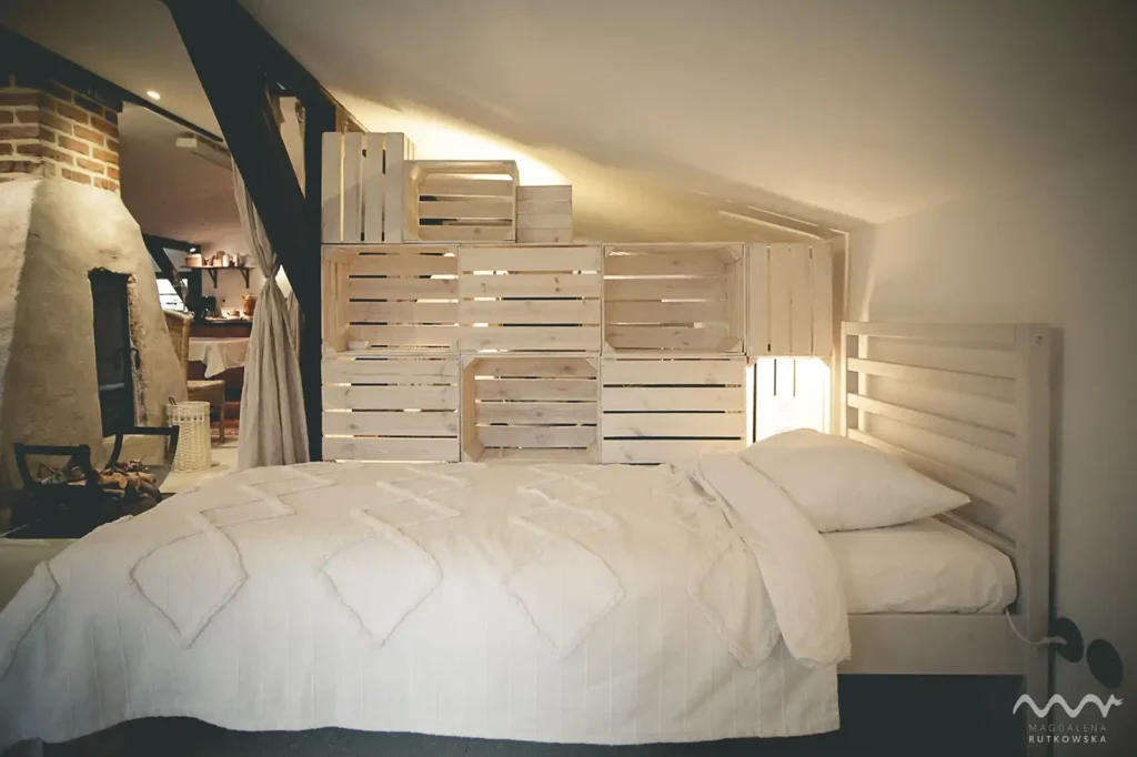 Poddasze, nocleg. Przytulne łóżko z jasną pościelą na Poddaszu w Łuby Siedem, na tle drewnianej, ażurowej ścianki. Widać lampkę nocną i fragment kominka.